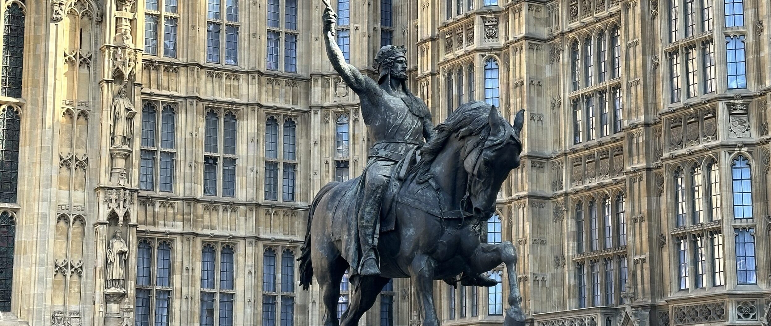 A bronze equestrian statue of a historical figure raising a sword stands proudly about an ornate, gothic-style building adorned with tall windows and intricate stonework.