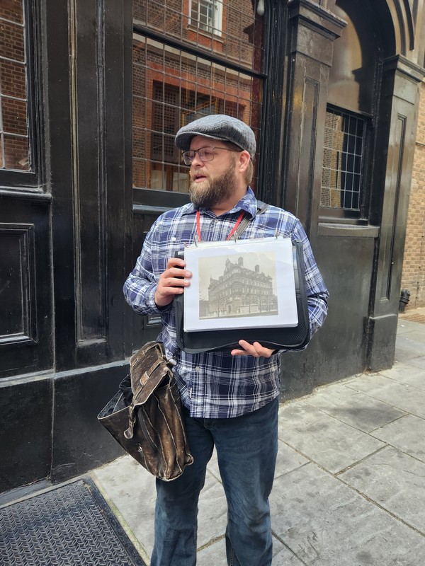 Wearing a cap and plaid shirt, guide Nate Loper holds a black folder featuring a historic black-and-white photo on a bustling London street. With a leather bag slung over his shoulder, he stands before a dark building facade, embarking on an enlightening Christian tour.