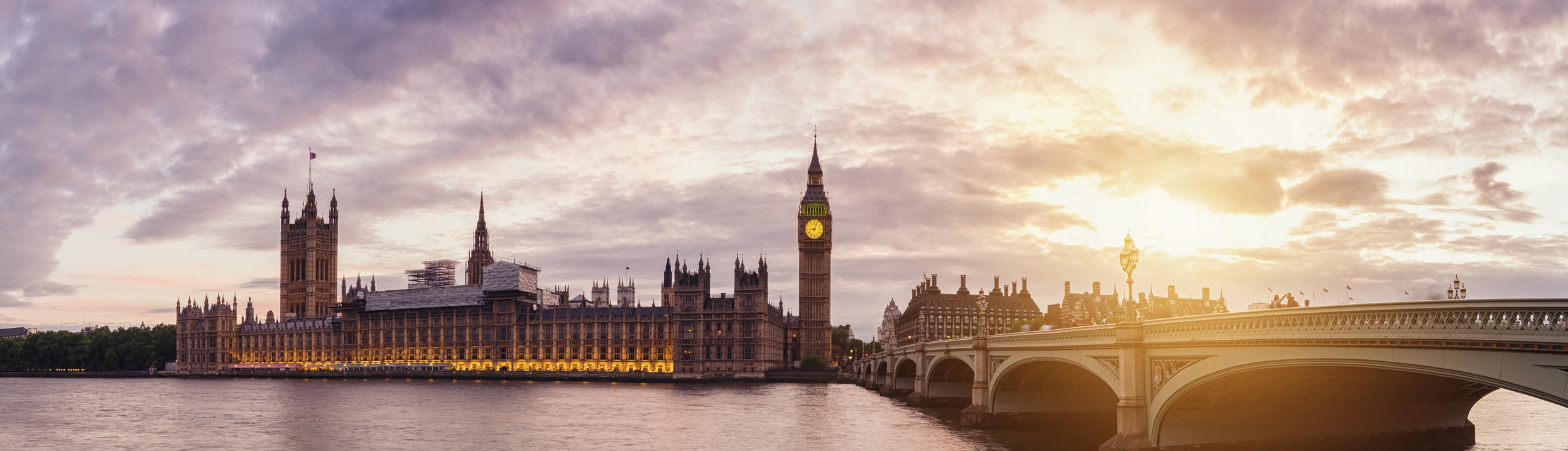 Panoramic view of the Houses of Parliament and Big Ben in London at sunset. The illuminated clock tower stands beside the River Thames, with Westminster Bridge on the right. The sky is partly cloudy with a warm glow.