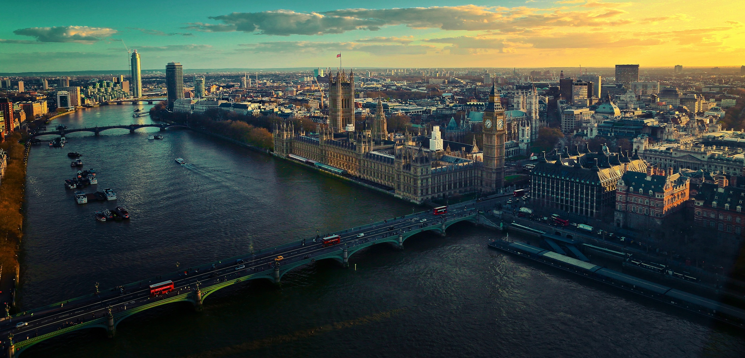 Aerial view of London at sunset, featuring the River Thames with several bridges. The Houses of Parliament and Big Ben stand proudly among city buildings. The sky is a blend of blue and orange hues. Red buses dot the roads, capturing quintessential London in every Trip FAQs dream image.