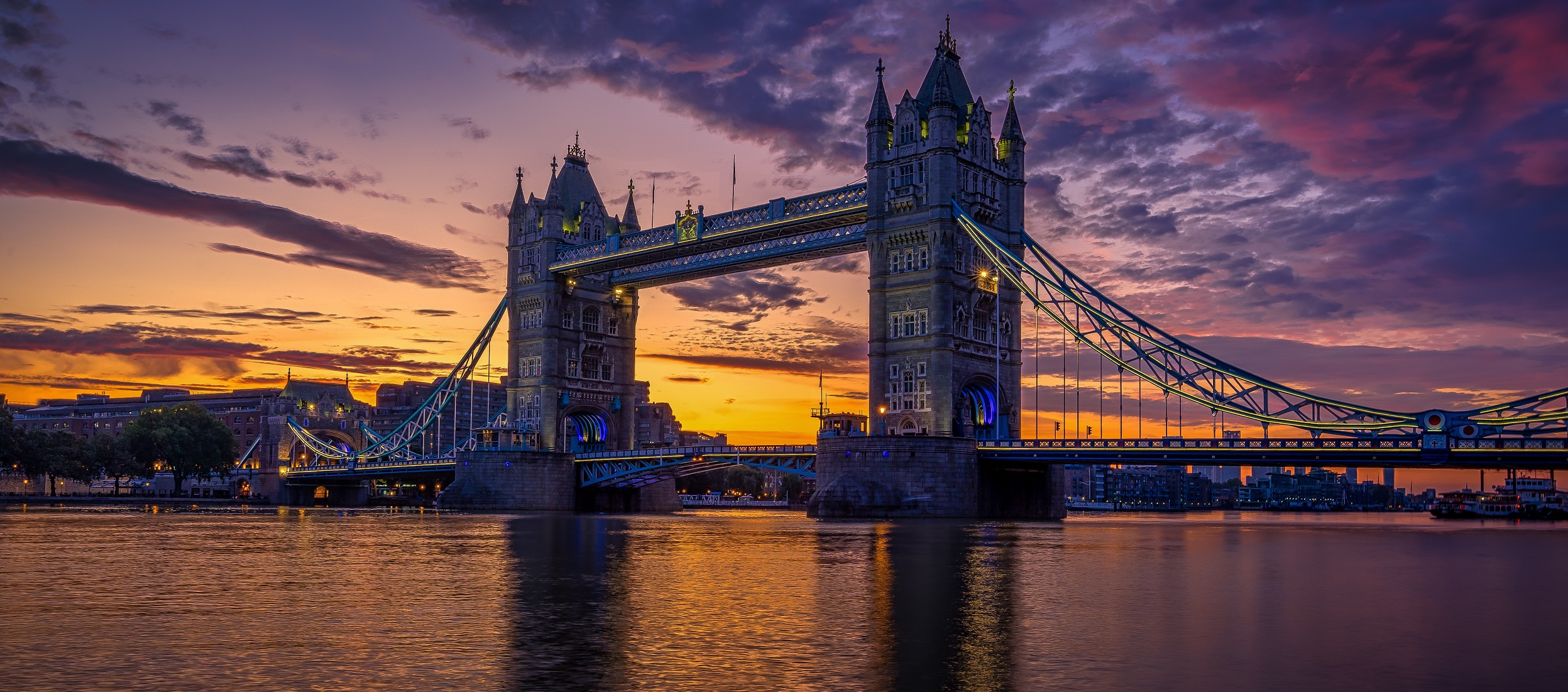 A stunning view of Tower Bridge in London during sunset, where the sky is painted with vibrant hues of orange, purple, and pink, reflecting off the calm river below. The bridges lights are illuminated, highlighting its iconic towers and structure—priceless compared to its construction cost.