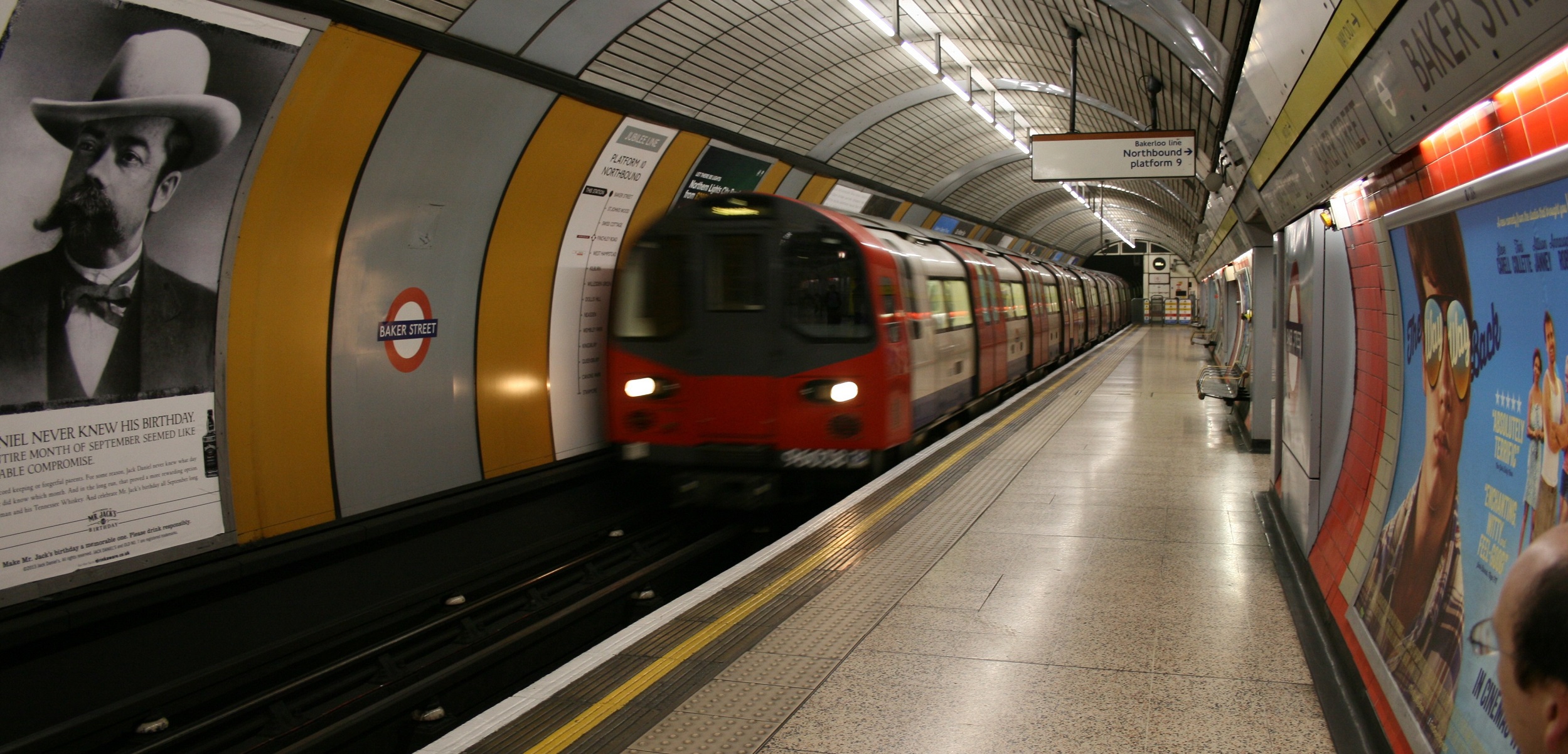 A London Underground train speeds through Baker Street station, where posters line the curved yellow and gray walls. The platform is mostly empty except for a few distant figures, while a Sherlock Holmes-themed poster invites you to book a London tour steeped in mystery and intrigue.