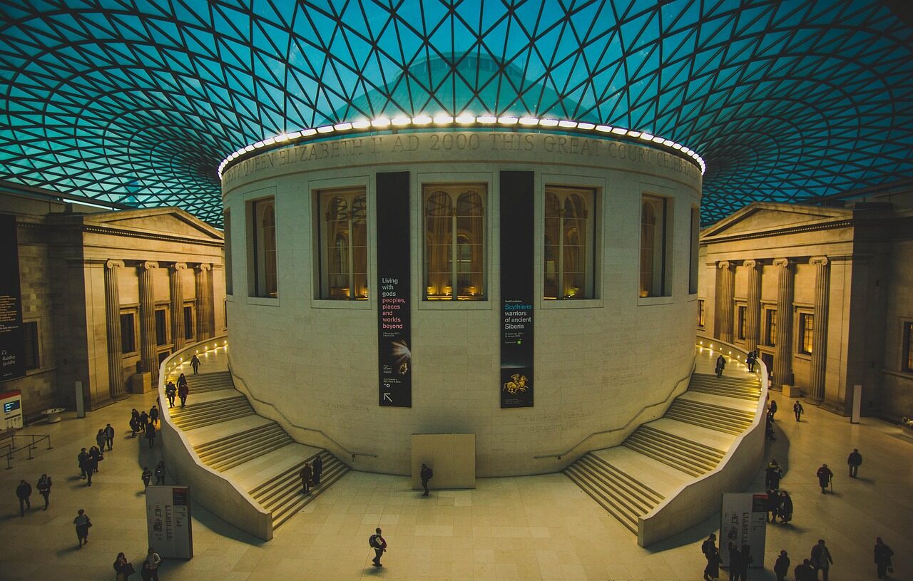 People walking inside a grand museum hall with a high, blue lattice roof are captivated by the rich history of London. The circular central structure is surrounded by staircases, while banners hang on the walls. Warm lighting complements the architectures columns, enhancing this cultural tour experience.