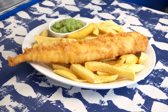 A plate of crispy battered fish atop golden fries, served with a side of mashed green peas, is placed on a blue and white patterned tablecloth. Its a classic meal that might even top your Trip FAQ list for must-try dishes!.