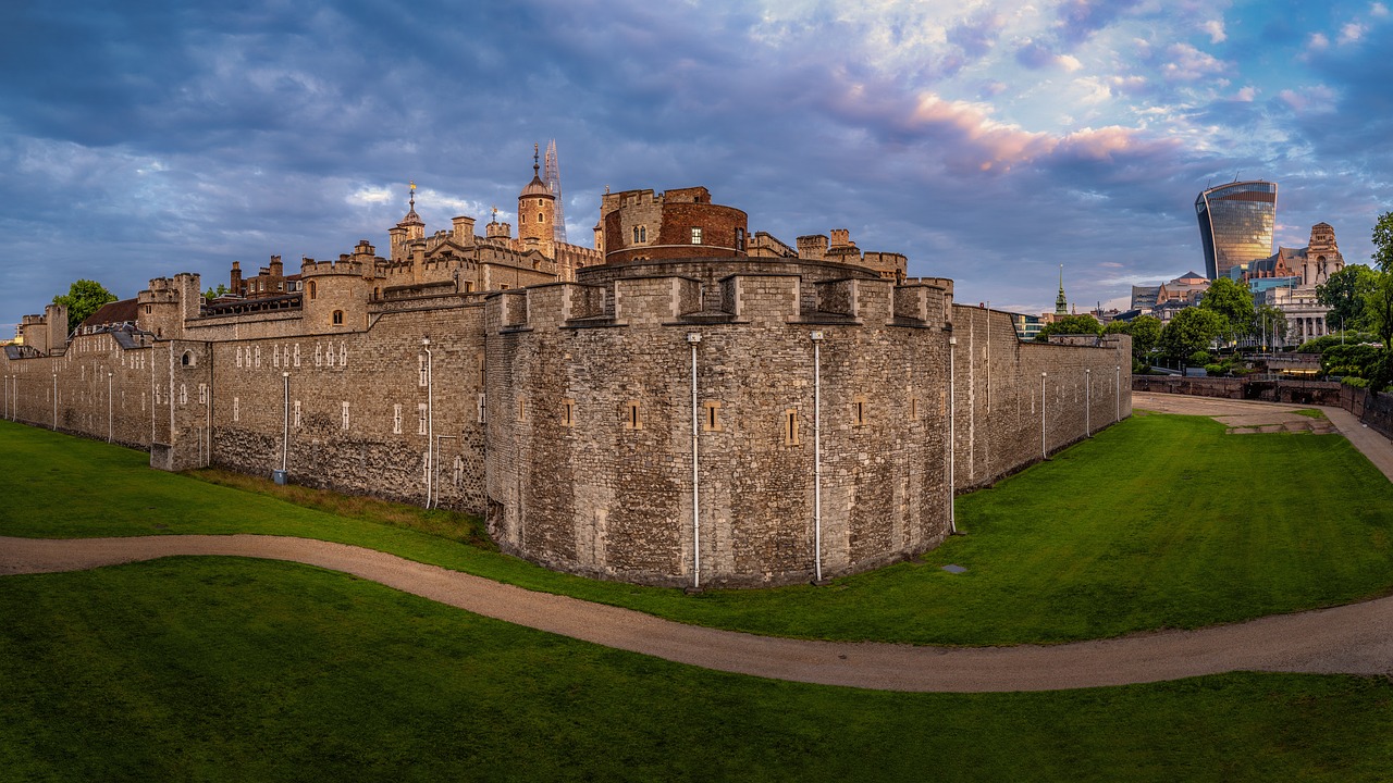 Panoramic view of the Tower of London, surrounded by lush lawns and a winding path. The historic stone structure stands in contrast to modern buildings under a cloudy sky, reminding visitors of its enduring legacy amid the citys evolving landscape and the priceless cost of history preserved.